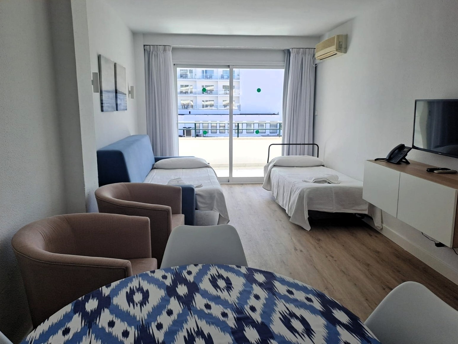 Una habitación luminosa y sencilla con dos camas individuales, un sofá azul, una mesa de comedor con sillas, un televisor y un balcón con vista a otros edificios.