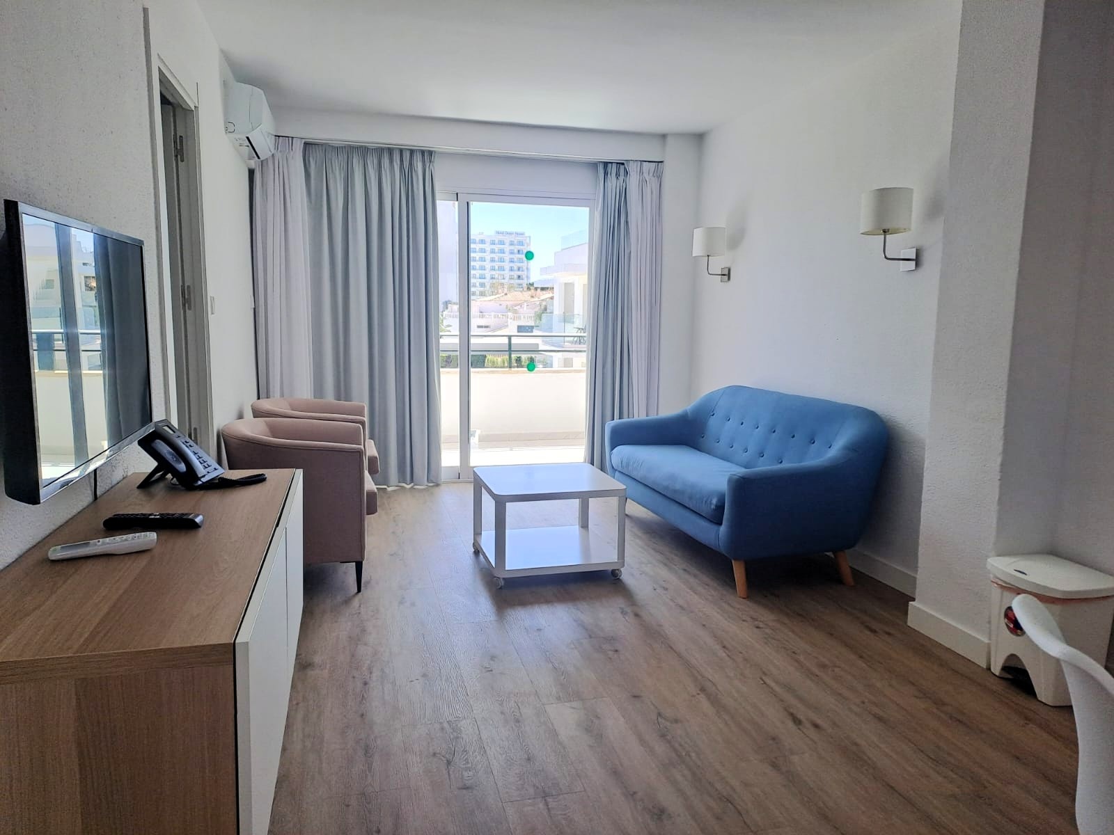 Una sala de estar luminosa presenta un sofá azul, dos sillones, un mueble con televisión, y un balcón al fondo.