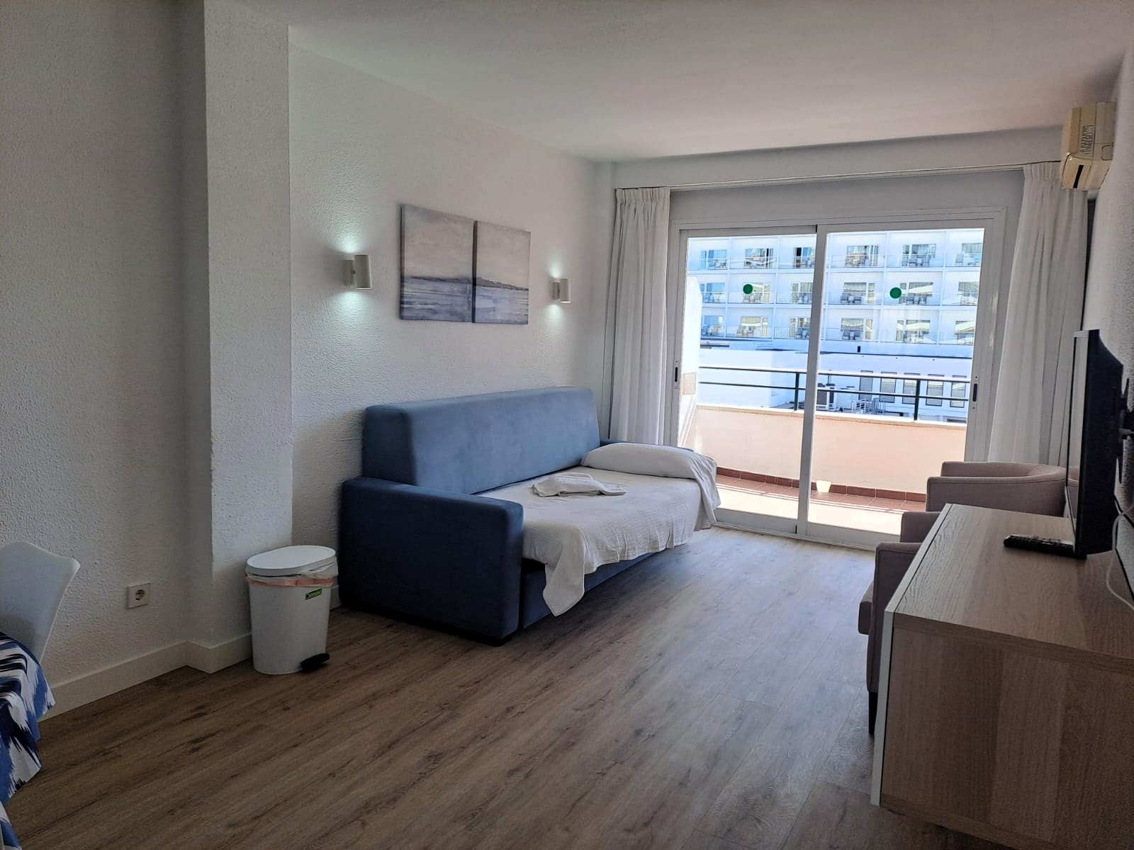 Un salón con sofá cama, una televisión y un balcón con vistas a edificios cercanos.