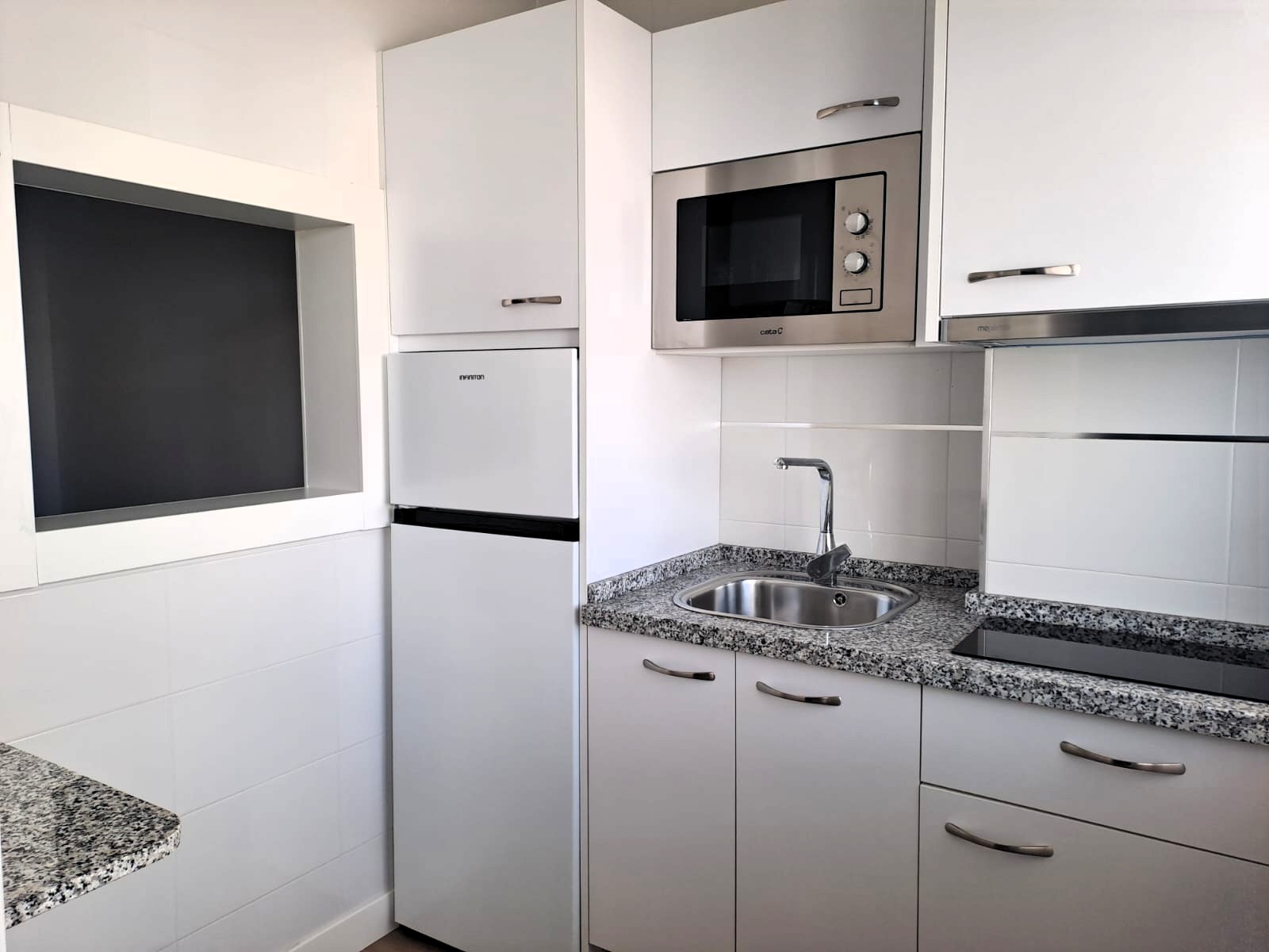 Una cocina moderna y compacta, predominantemente blanca, equipada con nevera, microondas integrado, fregadero de acero inoxidable y encimeras de granito.