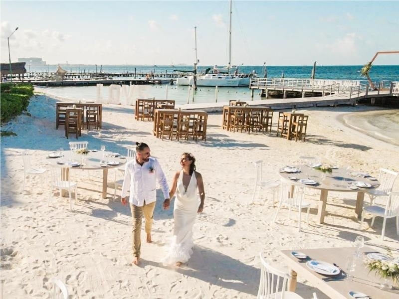 a bride and groom walking on a beach with tables and chairs