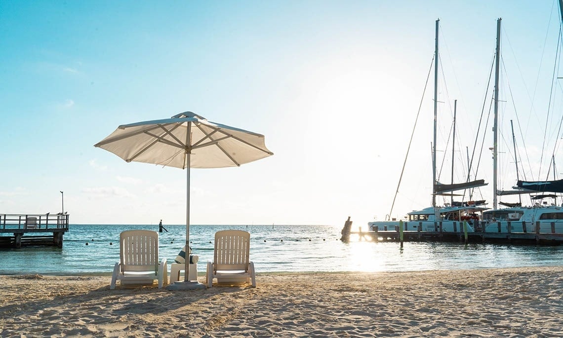 two chairs under an umbrella on a beach with boats in the background