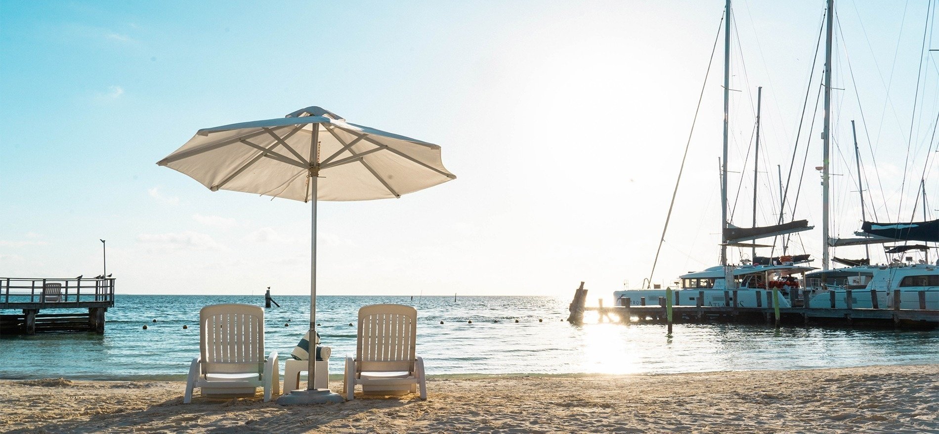 two chairs under an umbrella on a beach with boats in the background