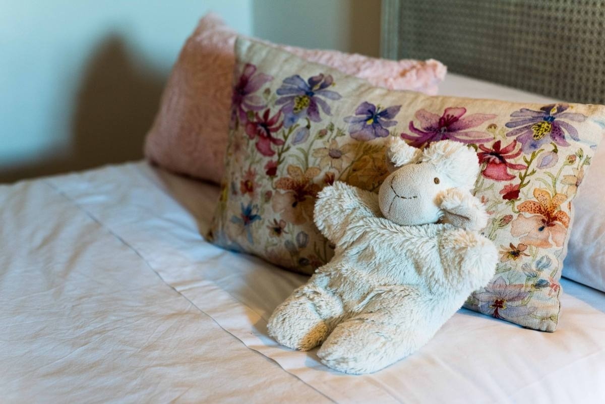 una oveja de peluche está sentada en una cama junto a una almohada floral