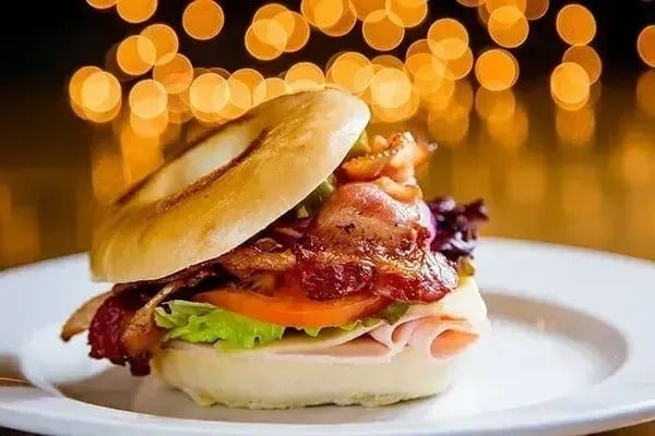 Auf einem weißen Teller steht ein Bagel-Sandwich mit Schinken , Salat , Tomaten und Speck .