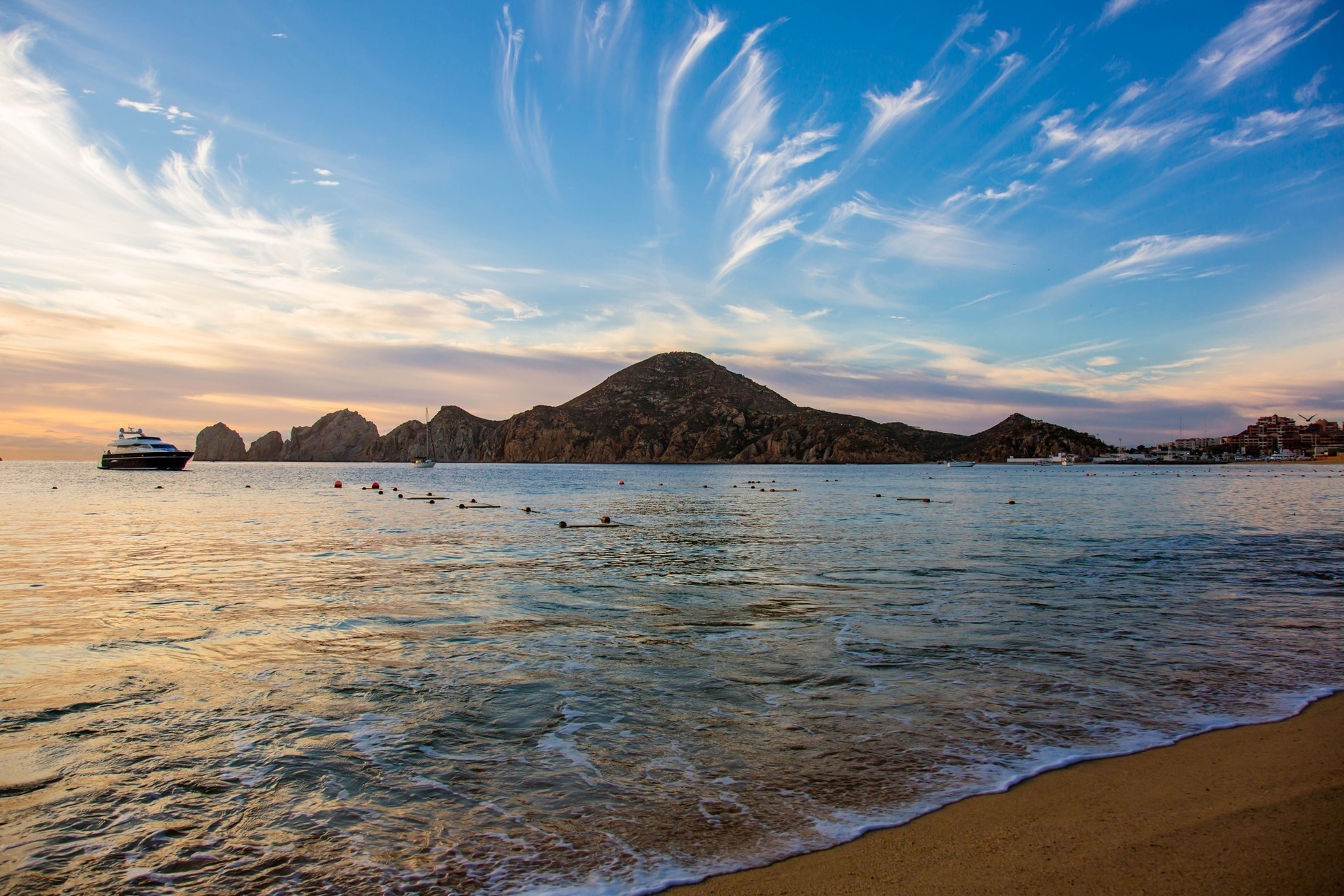 Un paisaje panorámico muestra un atardecer sobre el mar, destacando montañas rocosas, barcos y un cielo azul con nubes tenues y anaranjadas.