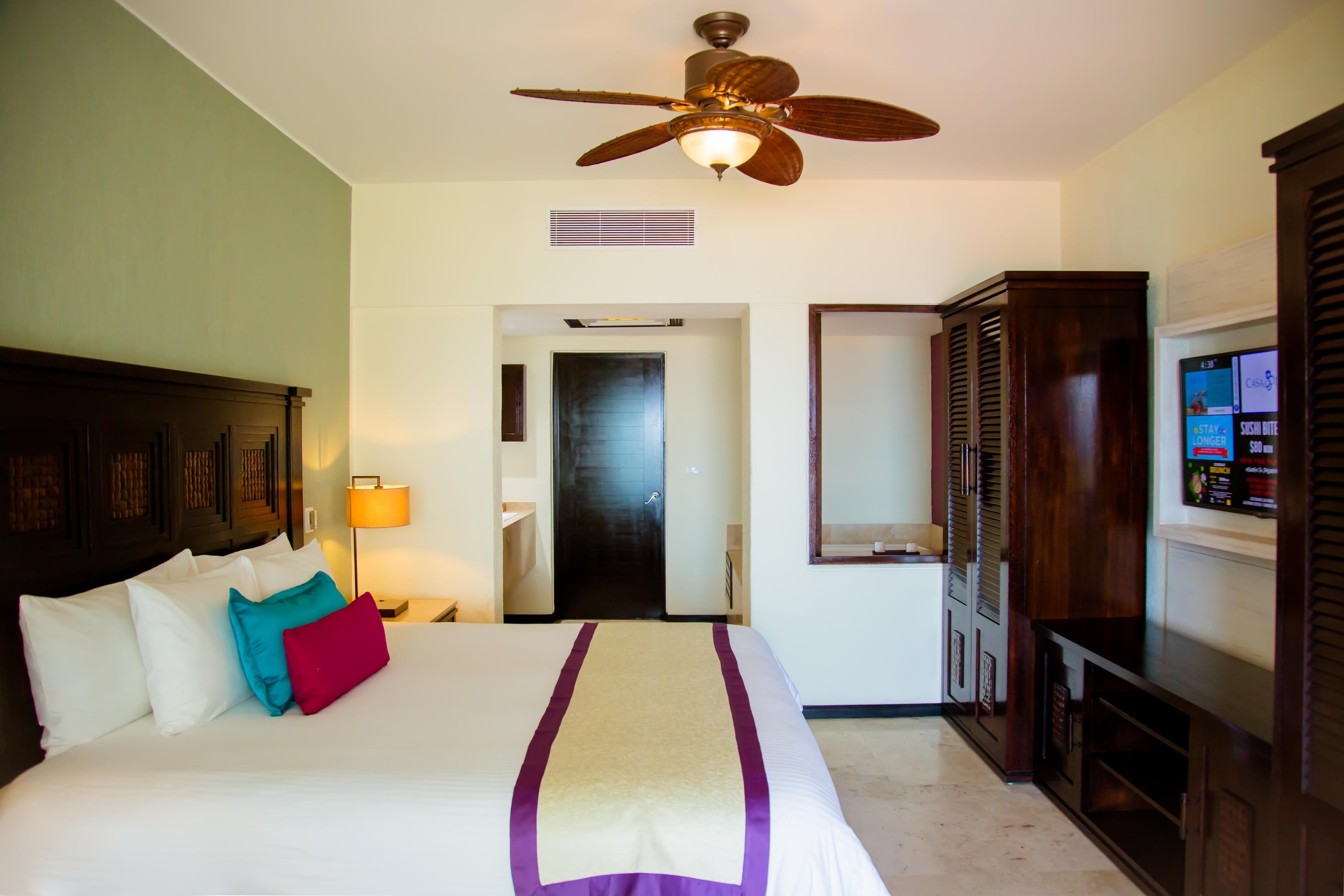 A well-appointed hotel room features a large bed with colorful accent pillows, a dark wood headboard, a ceiling fan, a flat-screen TV, a dark wood wardrobe, and an opening to a bathroom area with a sink and door.