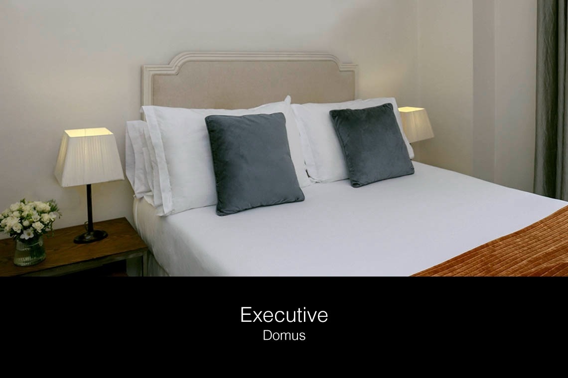 una cama con sábanas blancas y almohadas grises con la palabra ejecutiva domus al fondo