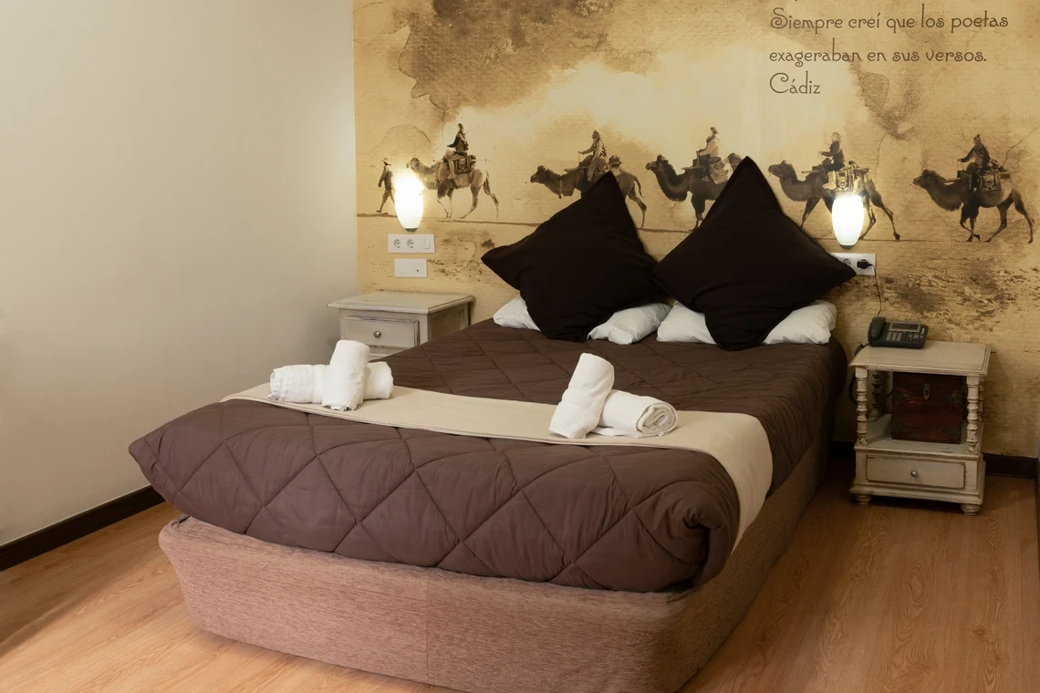 Cama doble con mural de desierto en Casual Alquimia Cádiz, mesitas de noche y decoración temática.