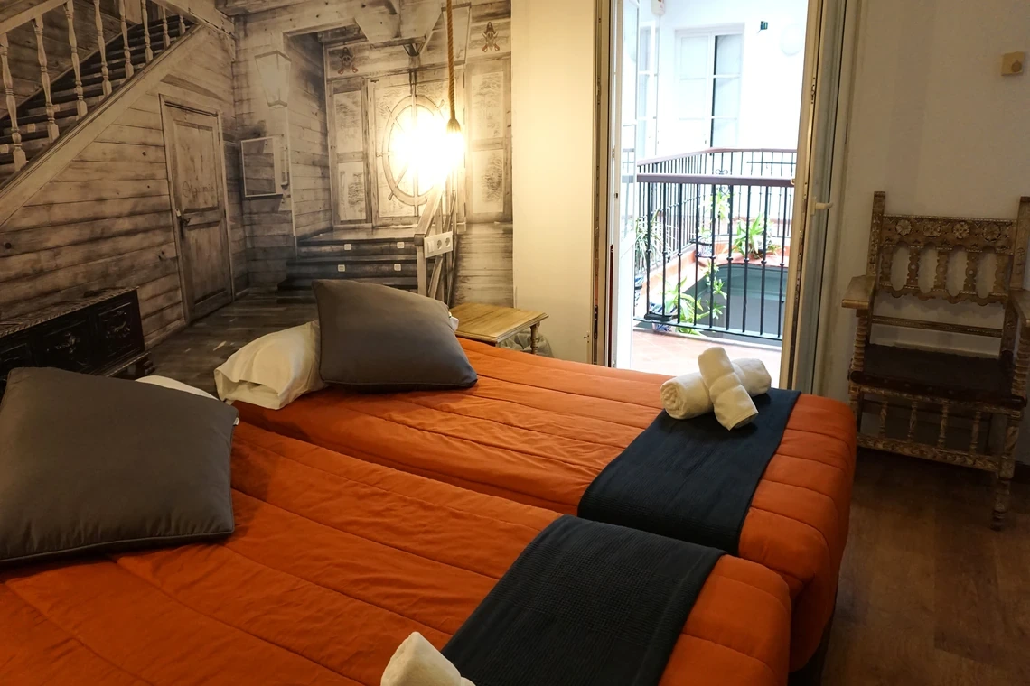 Habitación doble con dos camas naranjas, almohadas grises, y balcón exterior. Casual Alquimia Cádiz