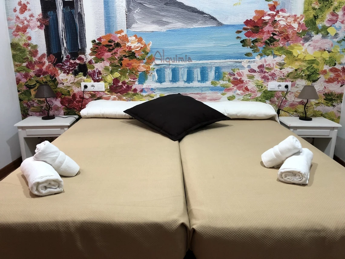 Cama doble acogedora con almohadas y toallas, mural floral y lámpara de mesita Casual Alquimia Cádiz.