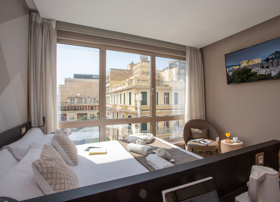 Modern hotel room with city view, large window, comfortable bed, and armchair. TV features Athens.
