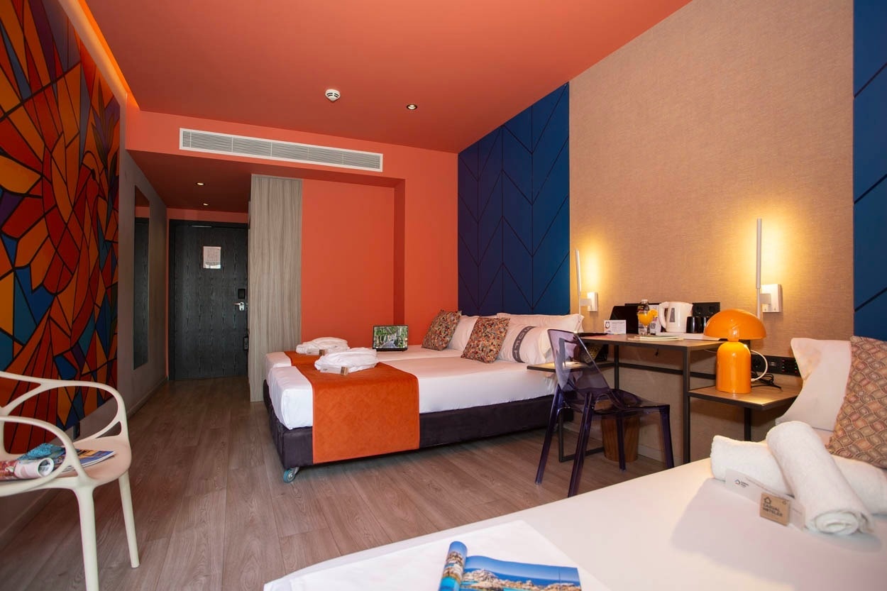 Modernes Hotelzimmer im Casual Kubic Athens – Doppelbett mit orangefarbener Tagesdecke, Schreibtisch und Kunst.