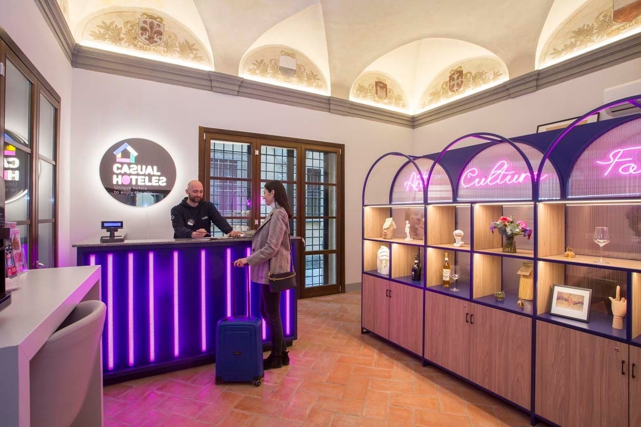 Casual Rinascimento Firenze: Empfangsbereich mit neonbeleuchtetem Tresen und charmanten Ausstellungsregalen eines zentralen Hotels mit Pool in Florenz.
