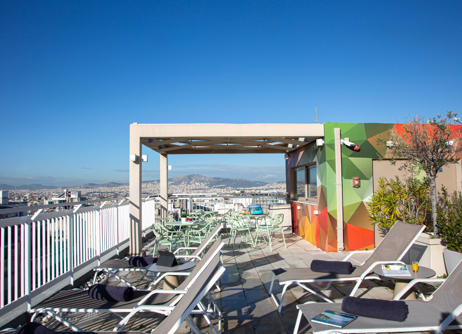 Hotel rooftop terrace with panoramic city views, lounge chairs, outdoor dining, and clear blue sky.