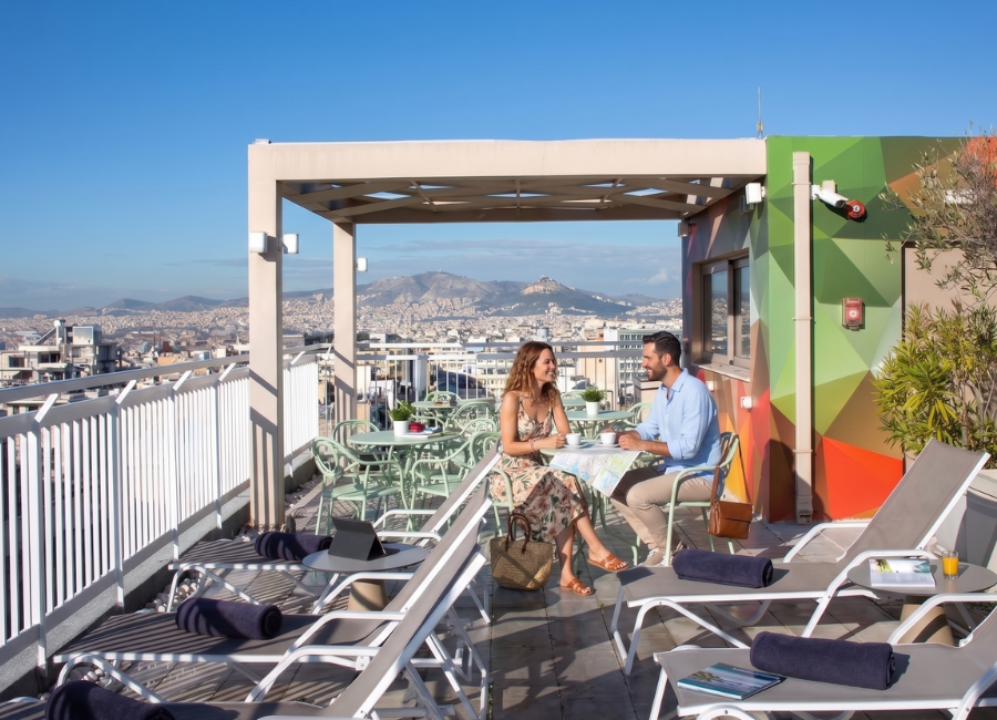 Hotel rooftop terrace with couple enjoying coffee and panoramic Athens city views, including Acropolis.
