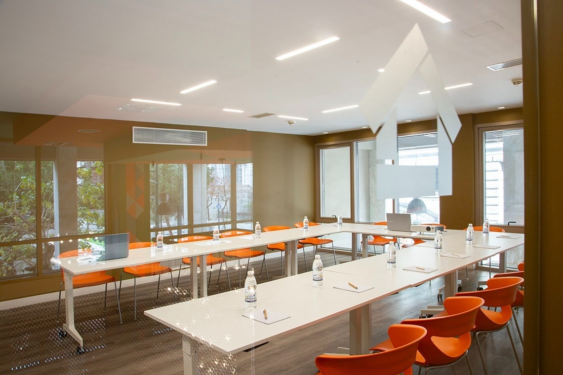 Una sala de reuniones moderna y luminosa con mesas dispuestas en forma de U, sillas de color naranja brillante y amplios ventanales.