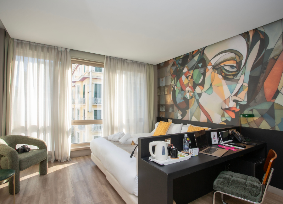 Stylish hotel room featuring a vibrant art mural, comfortable bed, work desk, and bright window.