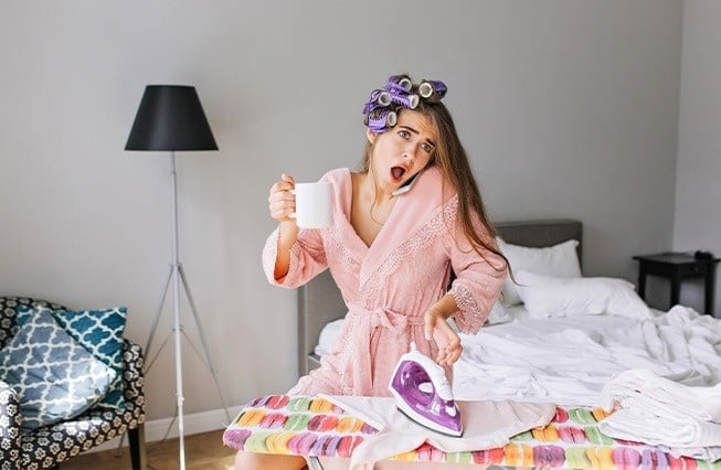 une femme portant des bigoudis parle sur un téléphone tout en tenant une tasse de café et un fer à repasser .