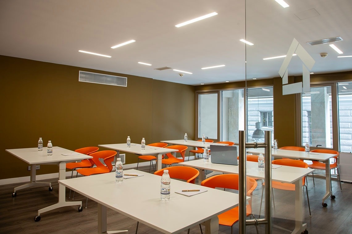 Una sala de reuniones o formación moderna y luminosa con paredes marrones, mesas blancas, sillas naranjas, botellas de agua y una puerta de cristal con un logotipo de casa.