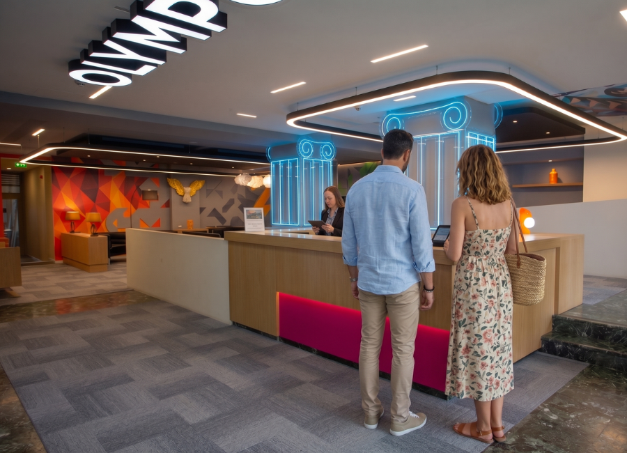 Guests check into a vibrant, modern hotel lobby with a stylish reception desk, bright neon accents, and 'OLYMP' sign.