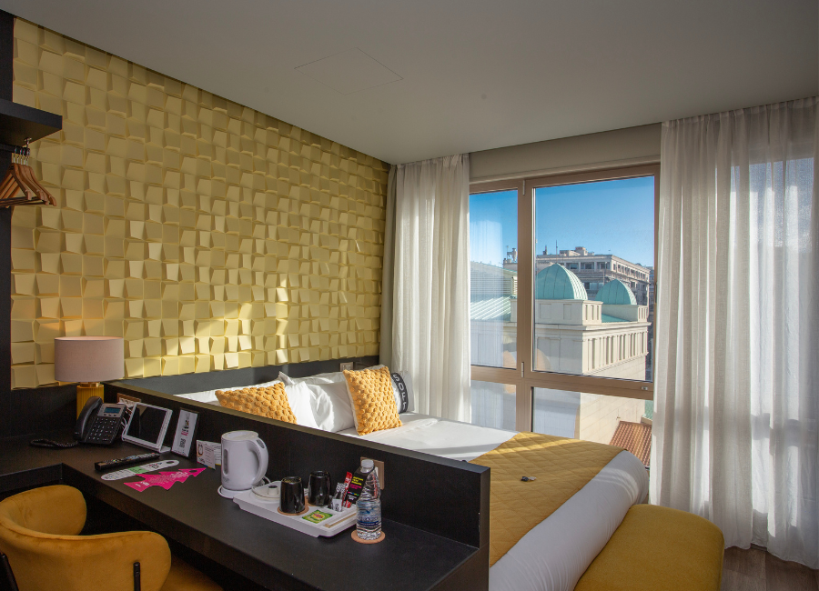Stylish hotel room with a golden textured accent wall, comfortable bed, desk, and large window with city views.