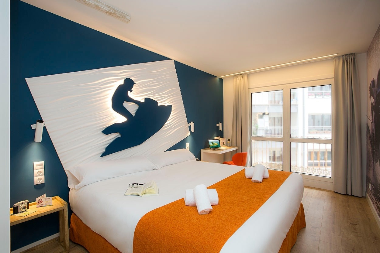 Geräumiges Schlafzimmer im Casual de las Olas San Sebastián: Doppelbett mit Surfer-Wandkunst, Schreibtisch und Fenster.