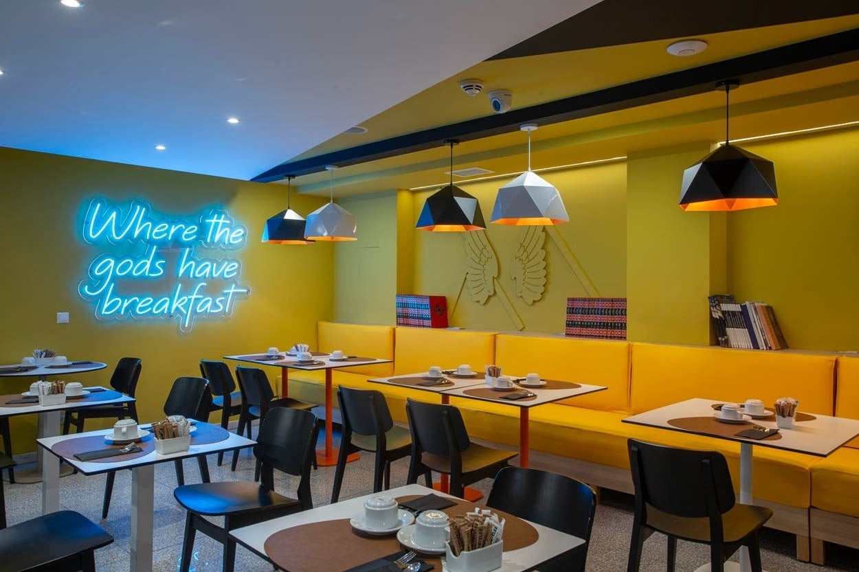 Frühstücksbereich des Casual Kubic Athens: Gelbe Sitzbänke & Neon-Zitat für einen lässigen Start in Athen.
