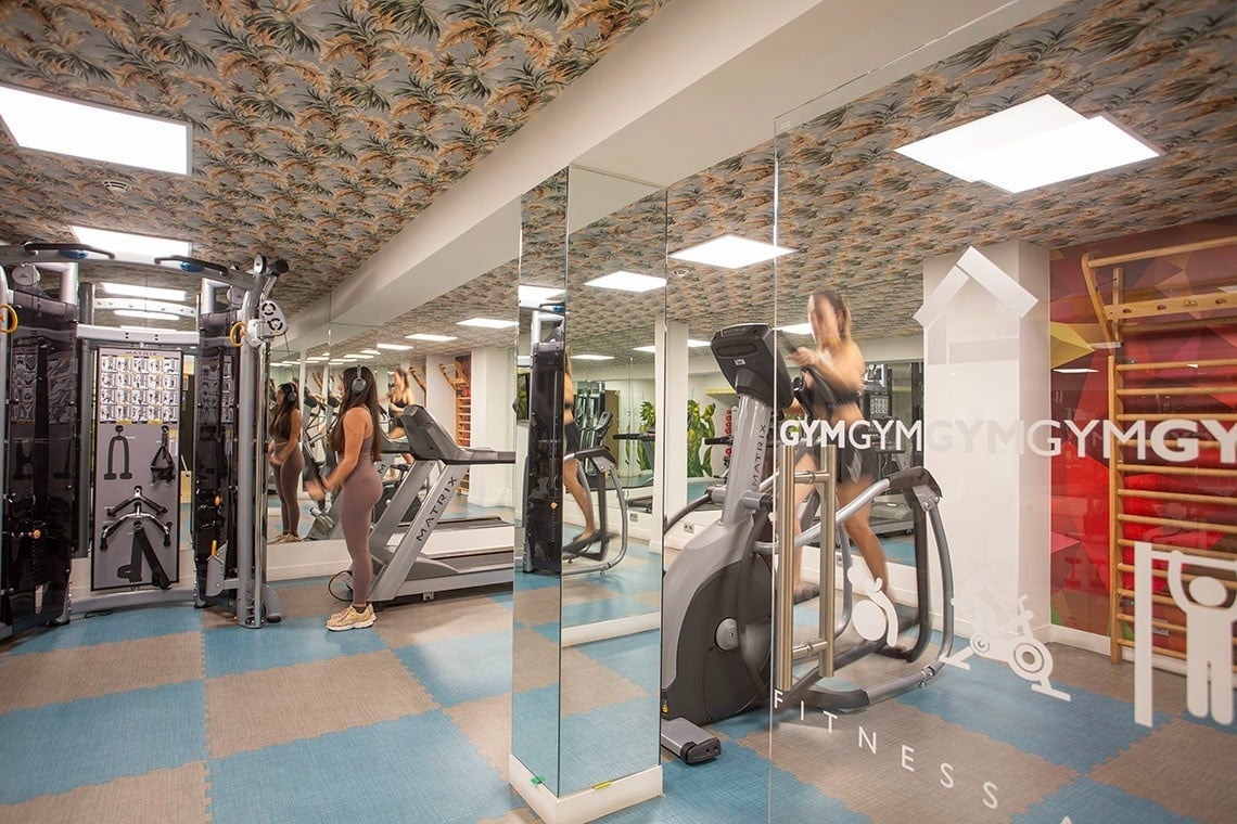 Un gimnasio moderno y luminoso con varias máquinas de ejercicio, dos mujeres entrenando, espejos en la pared y un techo con un estampado tropical.