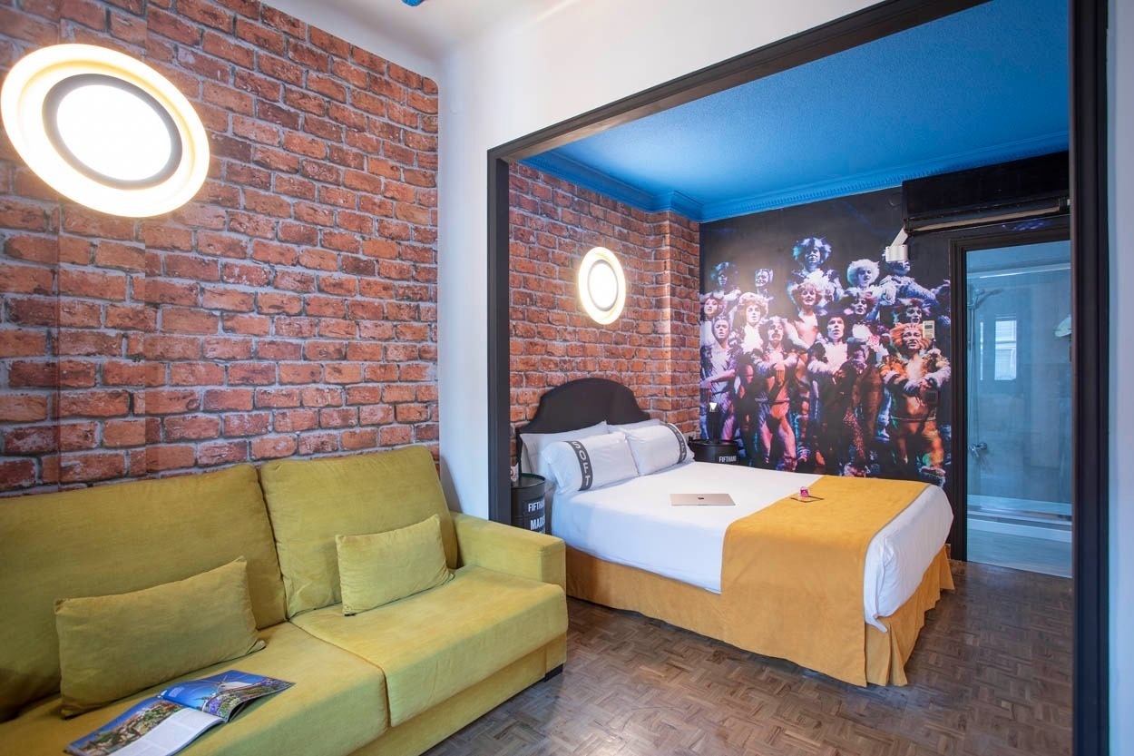 Ein lebhaft gestaltetes Zimmer mit einer gelben Couch, einer Ziegelsteinwand und einem Bett, das durch ein großes Wandbild des Musicals 