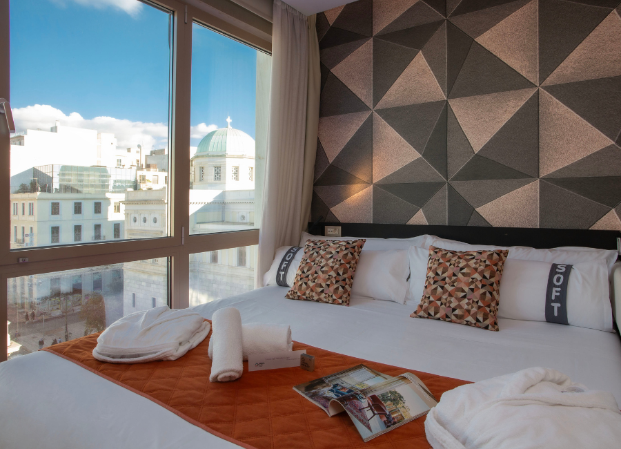 Bright hotel room with panoramic city view, comfy bed, soft towels, and modern geometric accent wall.