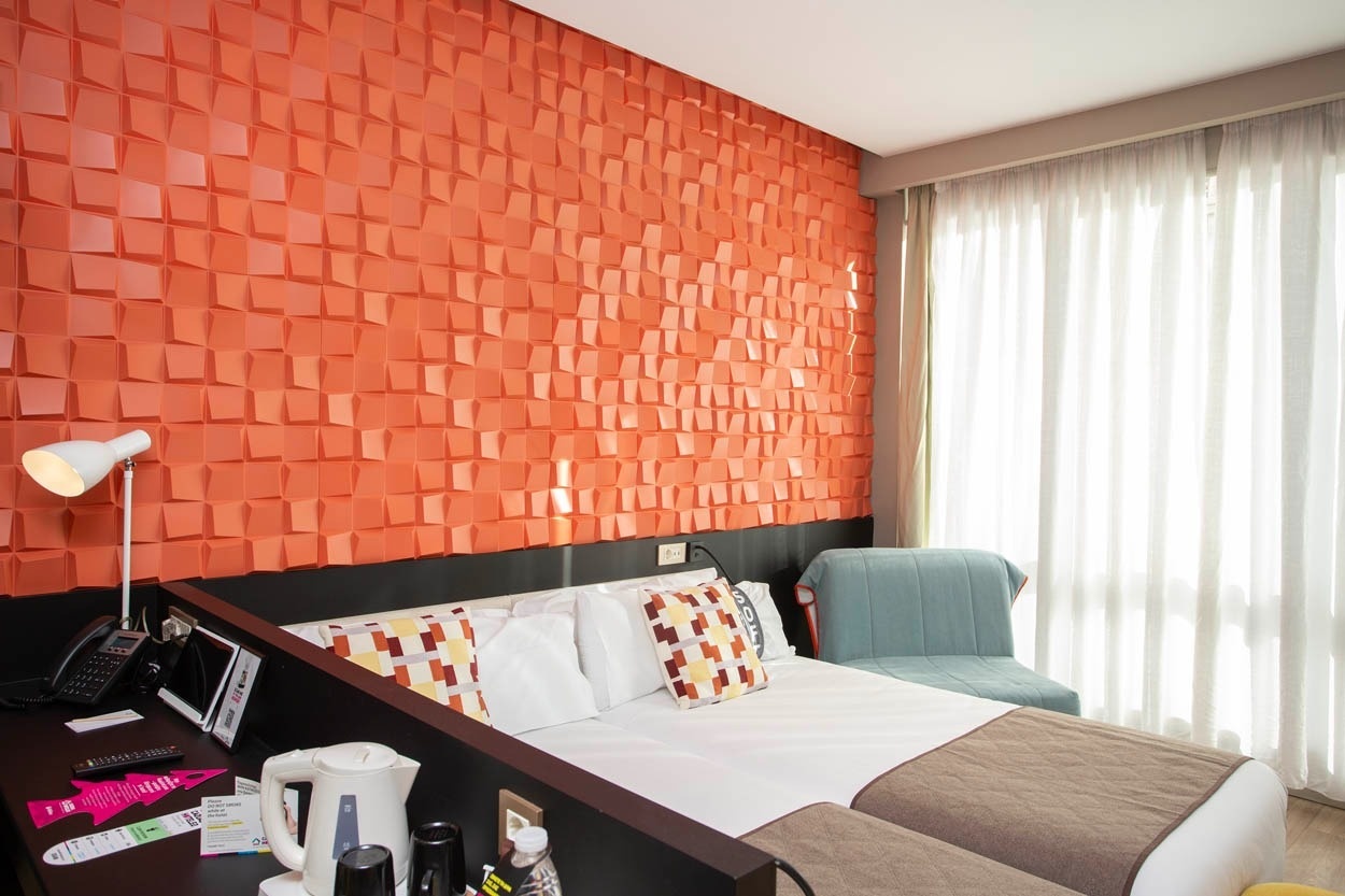 Modernes Hotelzimmer mit orangener 3D-Wand, Bett und Fenster. Casual Kubic Athens Hotel.