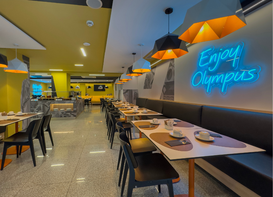 Modern hotel dining area with breakfast buffet, stylish seating, and 'Enjoy Olympus' neon sign.