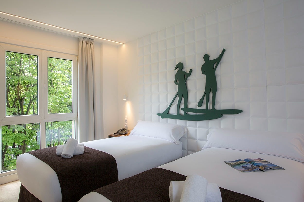 Modernes Hotelzimmer mit zwei Betten, Fenster mit Blick ins Grüne, Deko-Figuren. Casual de las Olas San Sebastián – Themenhotel.