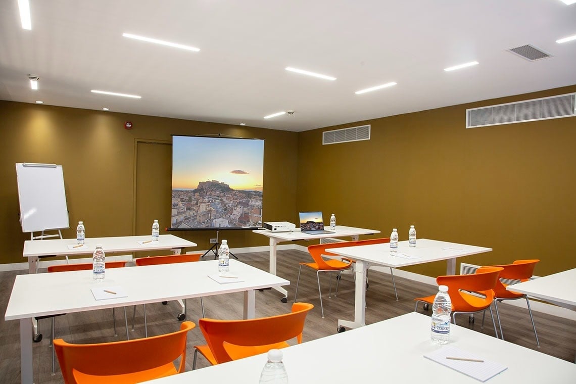 Una sala de reuniones moderna con mesas blancas, sillas naranjas, paredes marrón mostaza y una pantalla de proyector que muestra un paisaje urbano al atardecer.
