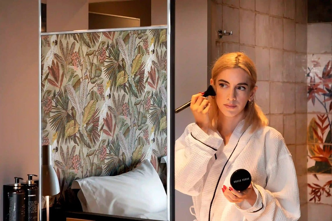 una mujer se aplica maquillaje frente a un espejo mientras sostiene una bolsa de bobbi brown