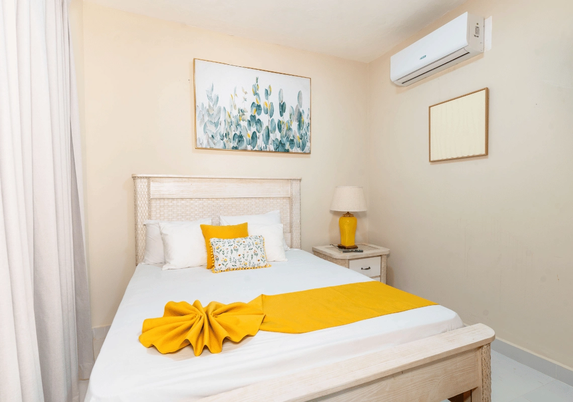 Cómoda cama con ropa de cama blanca y amarilla,Casual: Green Village Bayahibe junto a una ventana y una mesita de noche.