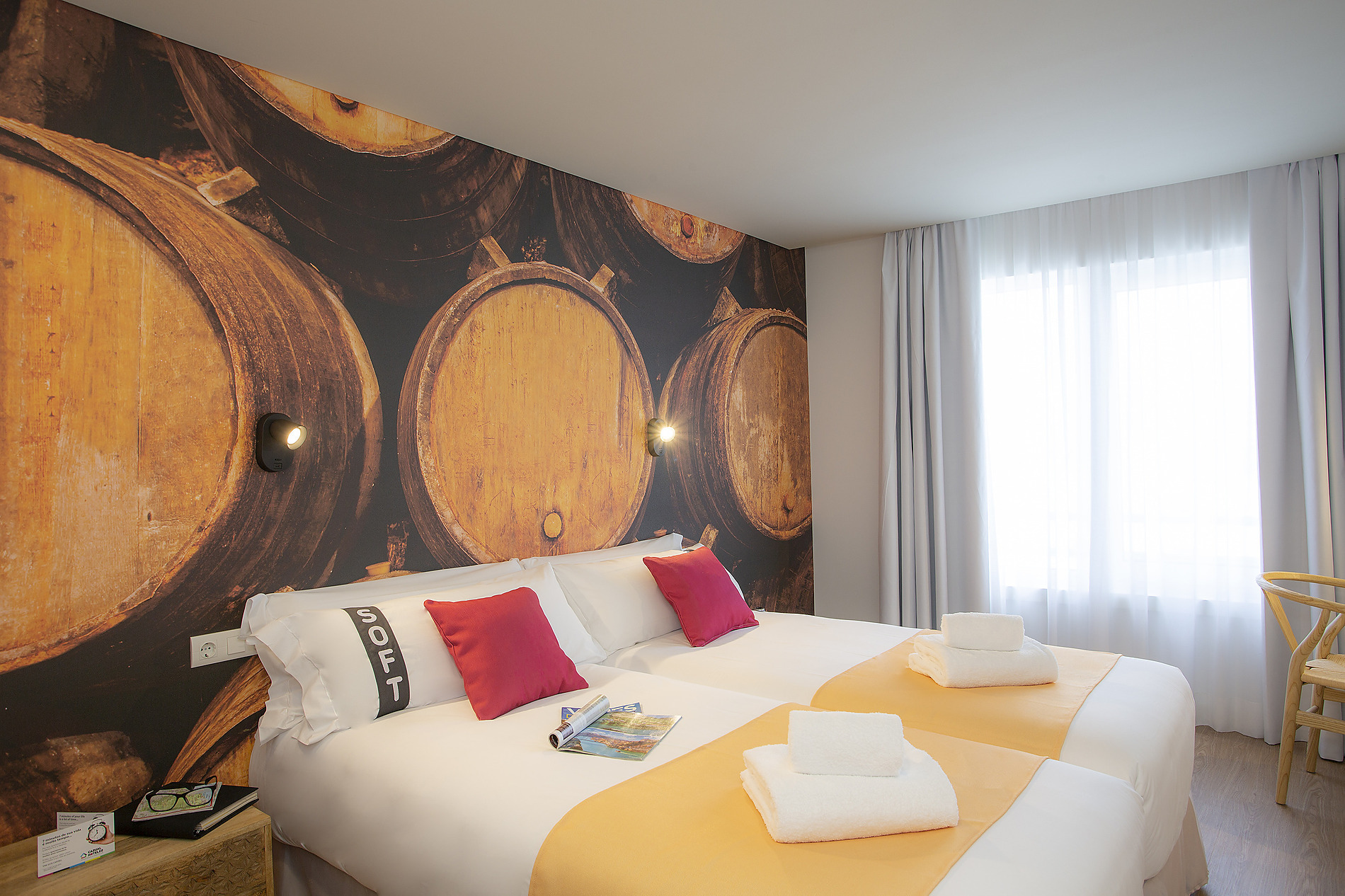 Cama doble temática de barricas de vino conCasual: Raizes Porto, almohadas rojas y blancas, toallas y cortinas claras.