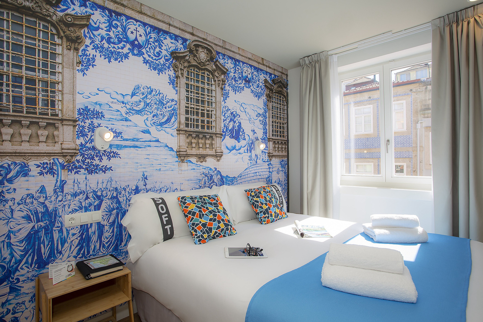 Habitación con mural estilo azulejo azul y blanco, cama acogedora con cojines vibrantes y ventana. Casual: Raizes Porto.