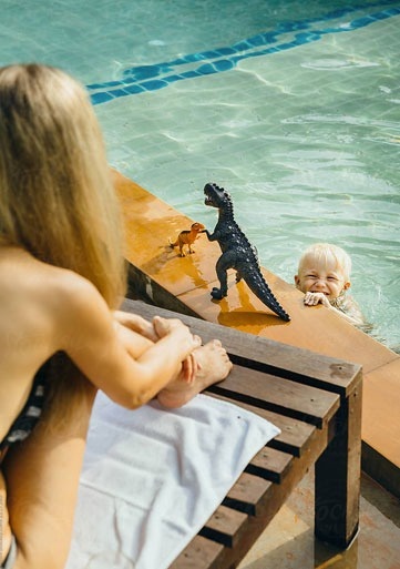 una mujer está sentada en un banco junto a una piscina mientras un niño juega con un dinosaurio de plástico .