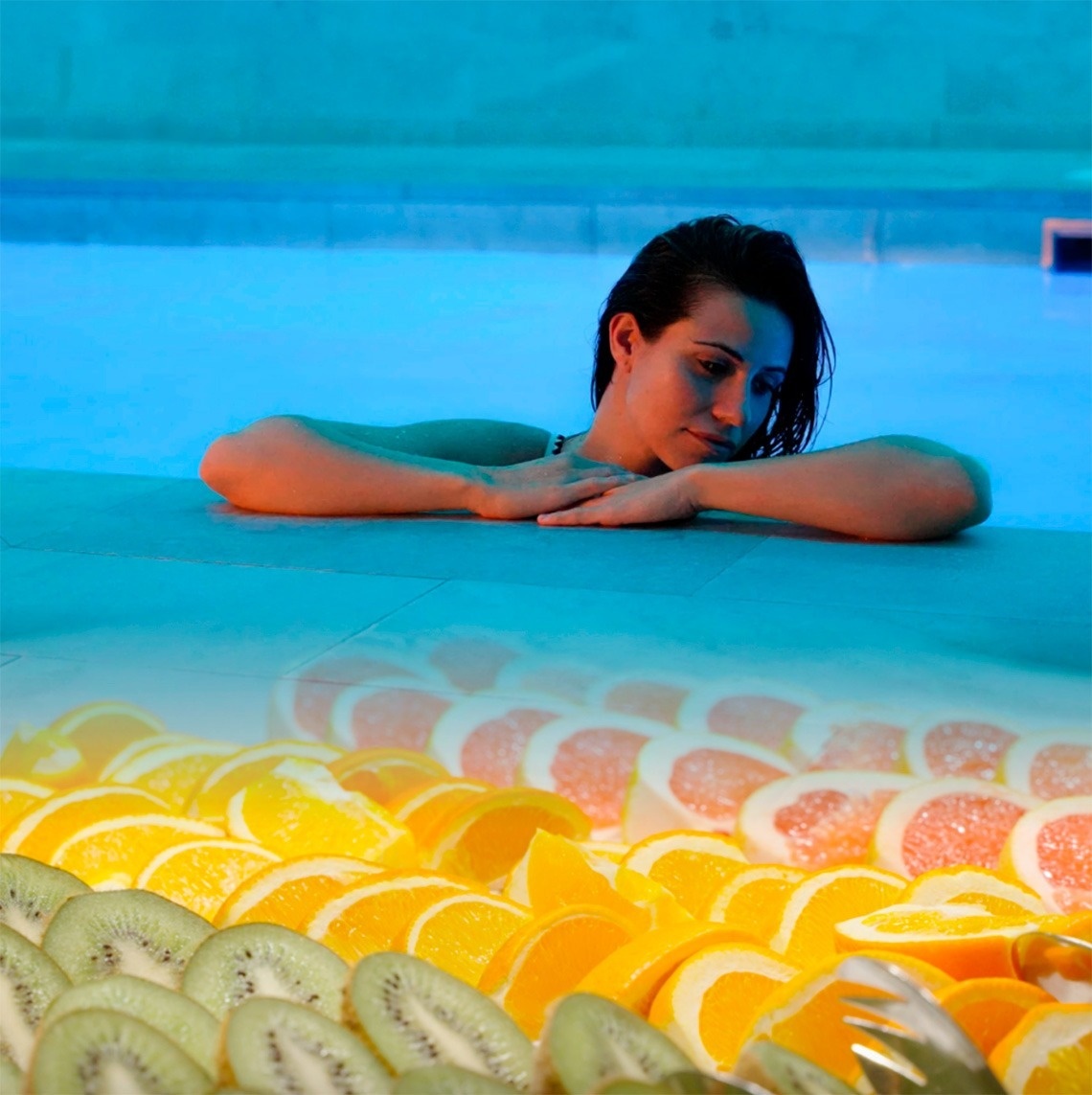 Una mujer se relaja en una piscina iluminada de azul, mientras la parte inferior de la imagen muestra una colorida disposición de kiwis, naranjas y pomelos en rodajas.