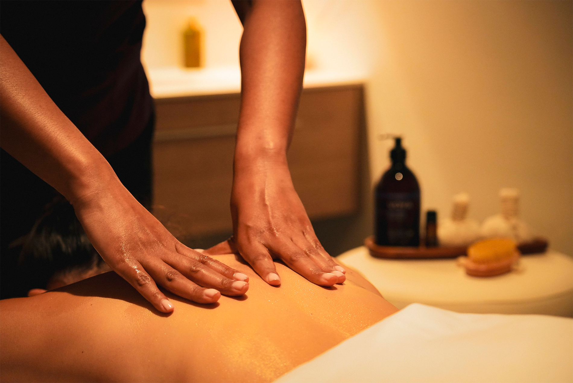 Una persona recibe un masaje de espalda con aceite en un ambiente de spa con productos de bienestar en el fondo.