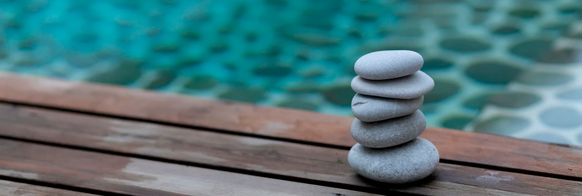 Pedras empilhadas em deck de madeira com piscina ao fundo, simbolizando desporto e bem-estar.