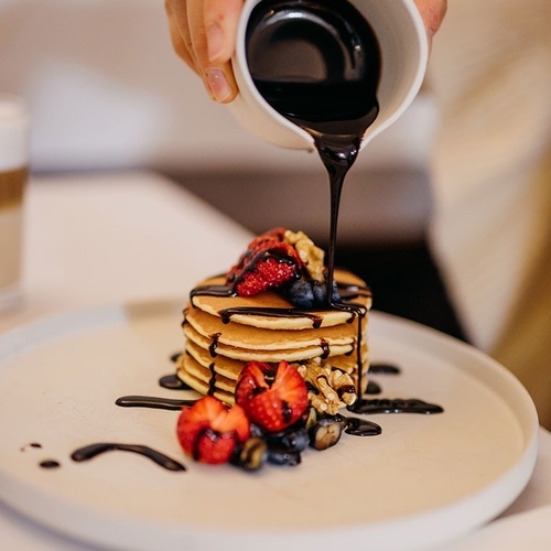 una persona vierte jarabe de chocolate sobre una pila de panqueques con bayas y nueces