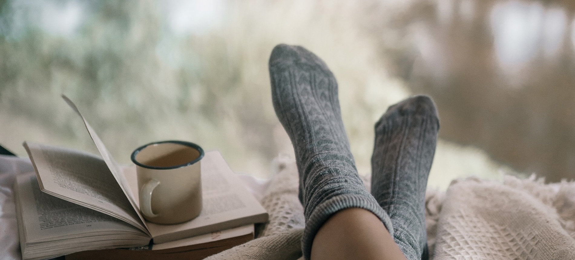 una persona con calcetines y una taza de café se sienta en una cama junto a un libro abierto