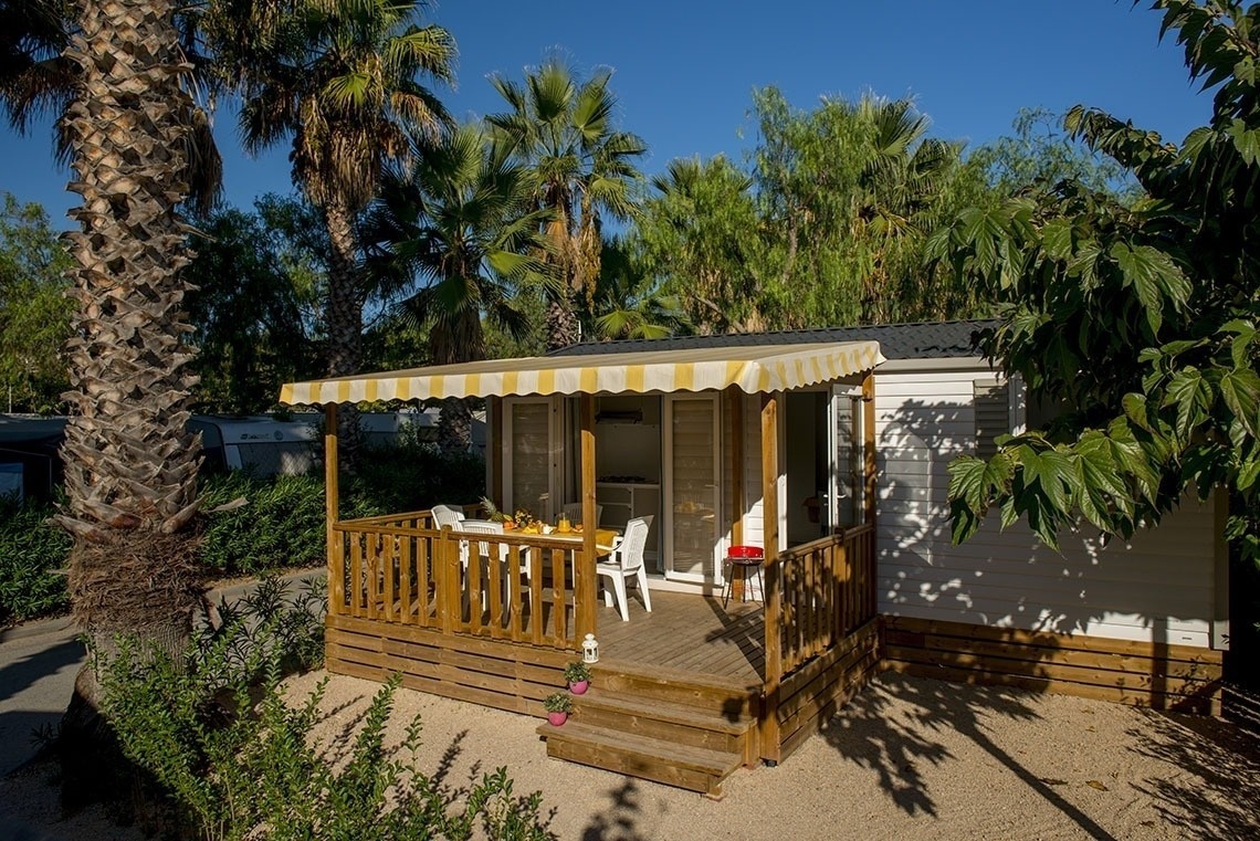 A cozy mobile home with a wooden deck, a yellow and white striped awning, and outdoor furniture is nestled among palm trees and lush greenery under a clear blue sky.