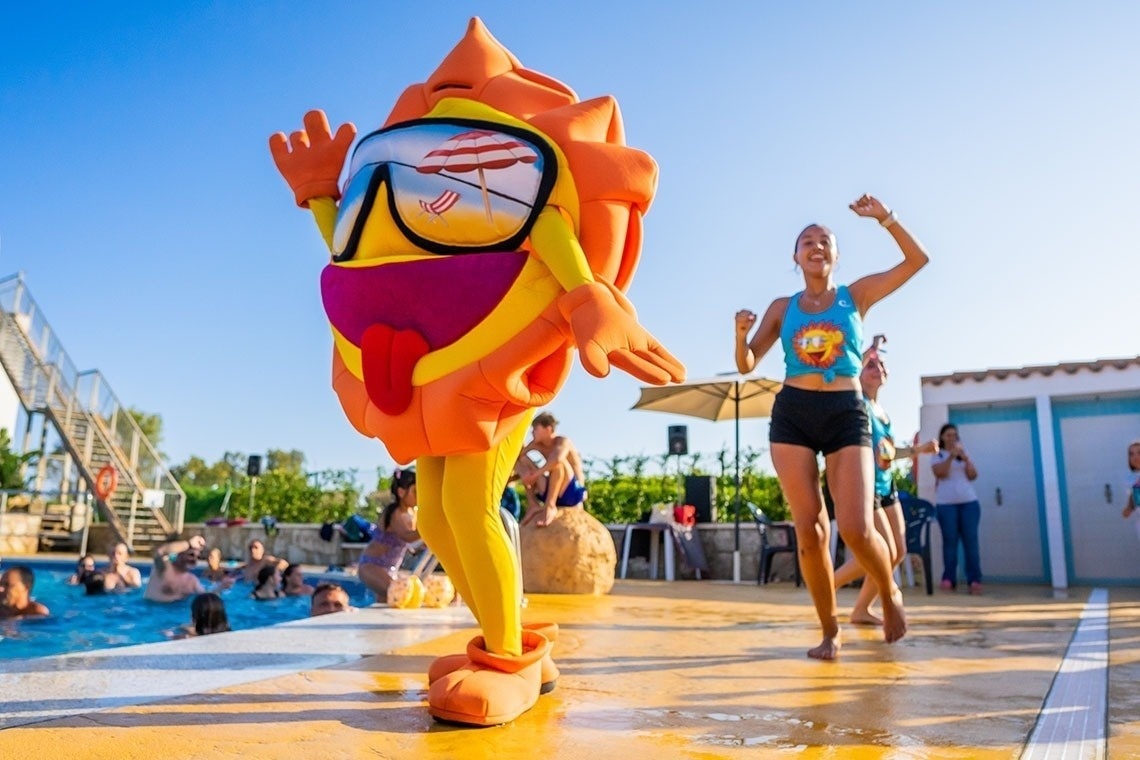 A person in a large, colorful sun mascot costume and an energetic woman dance by a swimming pool, filled with people, on a sunny day.