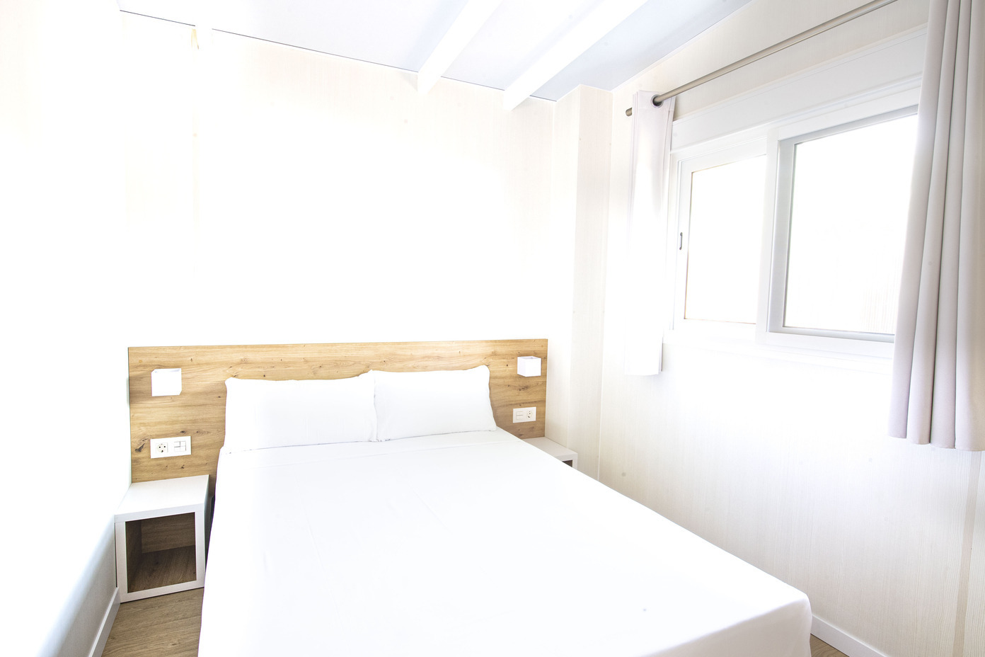Chambre d'hôtel moderne avec lit blanc, table de chevet et fenêtre. Un endroit parfait pour votre Creixell camping & family resort.