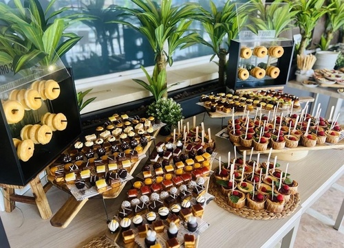 Una mesa presenta una abundante y elegante exhibición de postres variados, incluyendo donas, mini tartas y pastelitos, en un entorno iluminado con plantas de fondo.