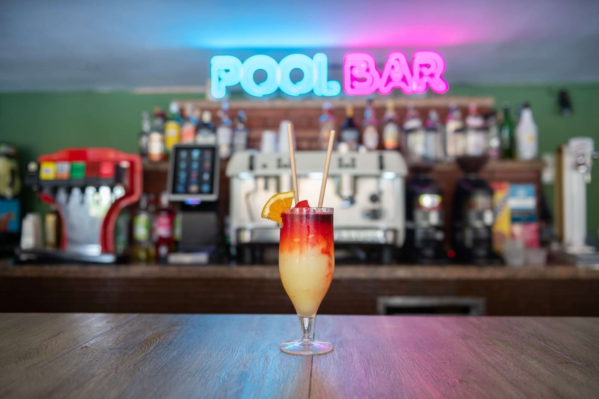 Un cóctel vibrante con guarnición de naranja y pajitas se apoya sobre una barra de madera, iluminado por un letrero de neón que dice "POOL BAR" en el fondo.